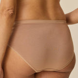 Rear view of a model wearing high-waisted seamless underwear in a light tan shade, paired with a matching bralette, against a solid beige background.