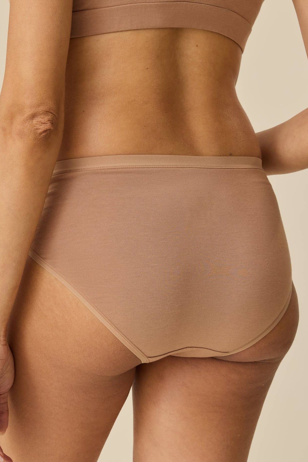 Rear view of a model wearing high-waisted seamless underwear in a light tan shade, paired with a matching bralette, against a solid beige background.