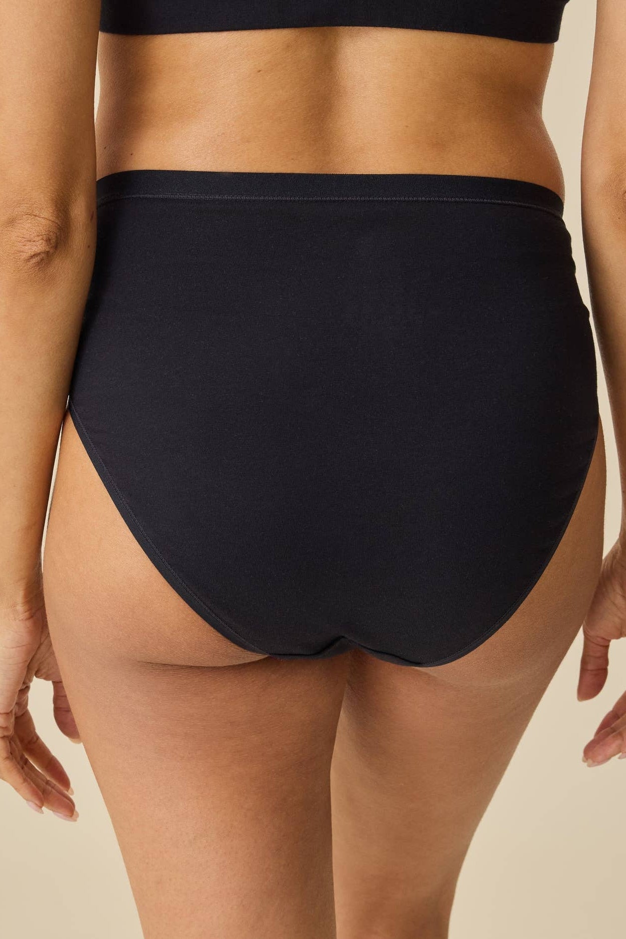 Rear view of a model wearing high-waisted black bikini bottoms against a plain beige background.