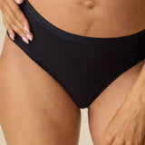 A close-up of a pregnant model wearing black maternity underwear, showing the waistband and hip coverage against a neutral beige background.