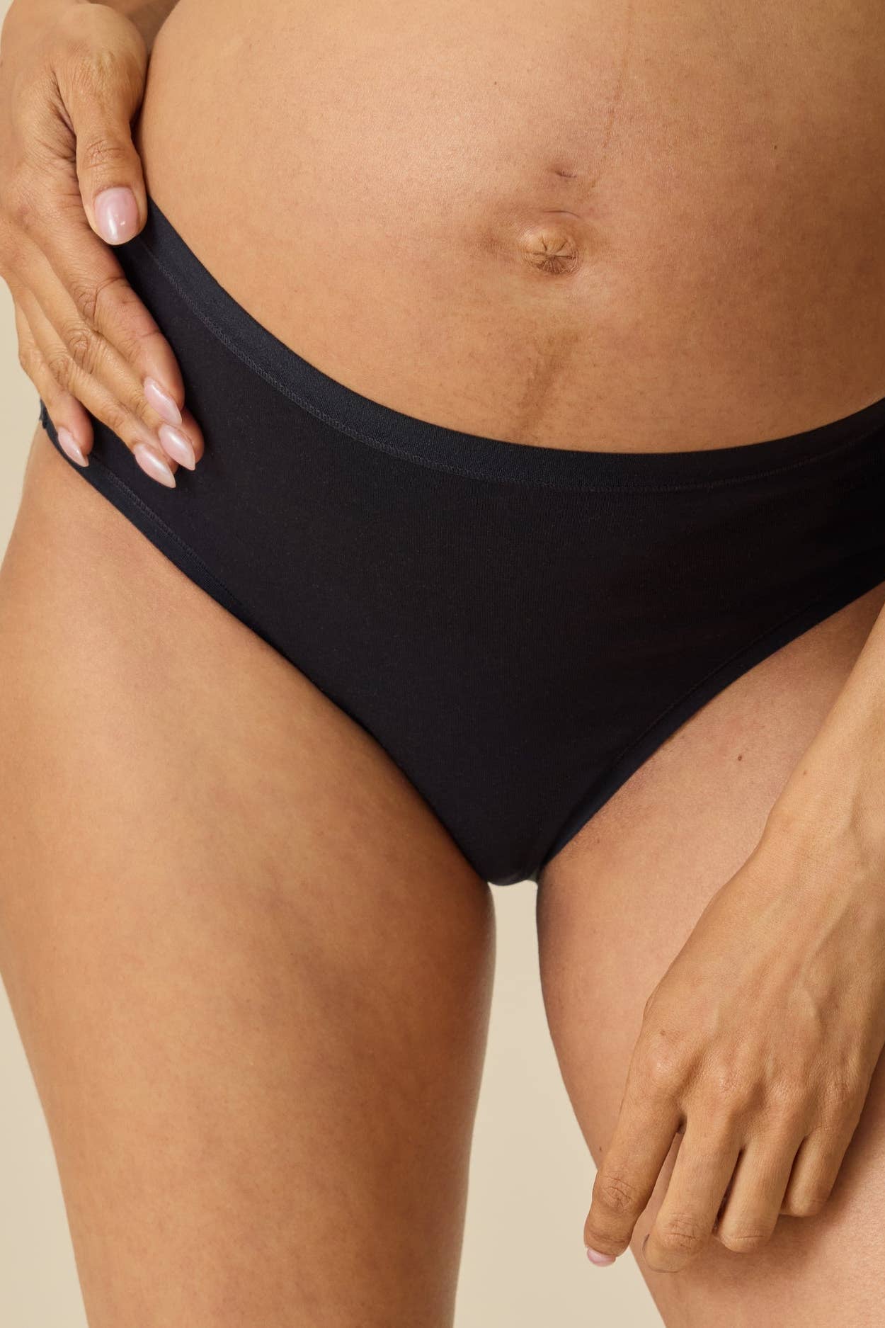 A close-up of a pregnant model wearing black maternity underwear, showing the waistband and hip coverage against a neutral beige background.