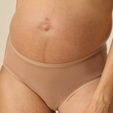 A close-up of a pregnant model wearing high-waisted, seamless maternity underwear in a warm tan shade, paired with a matching bralette.
